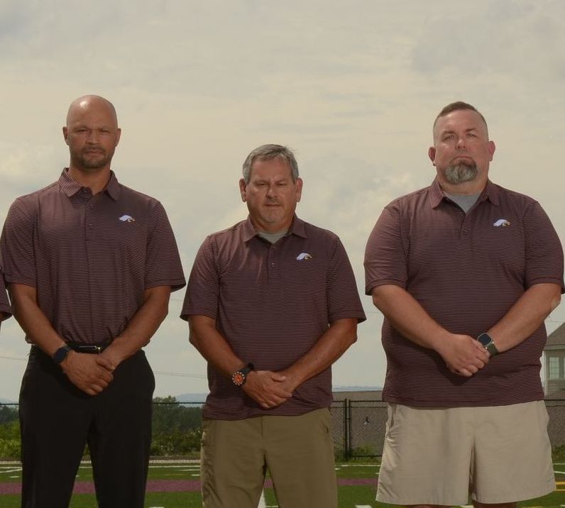Three men in maroon shirts and shorts/pants stand on a sports field, looking down with closed eyes.