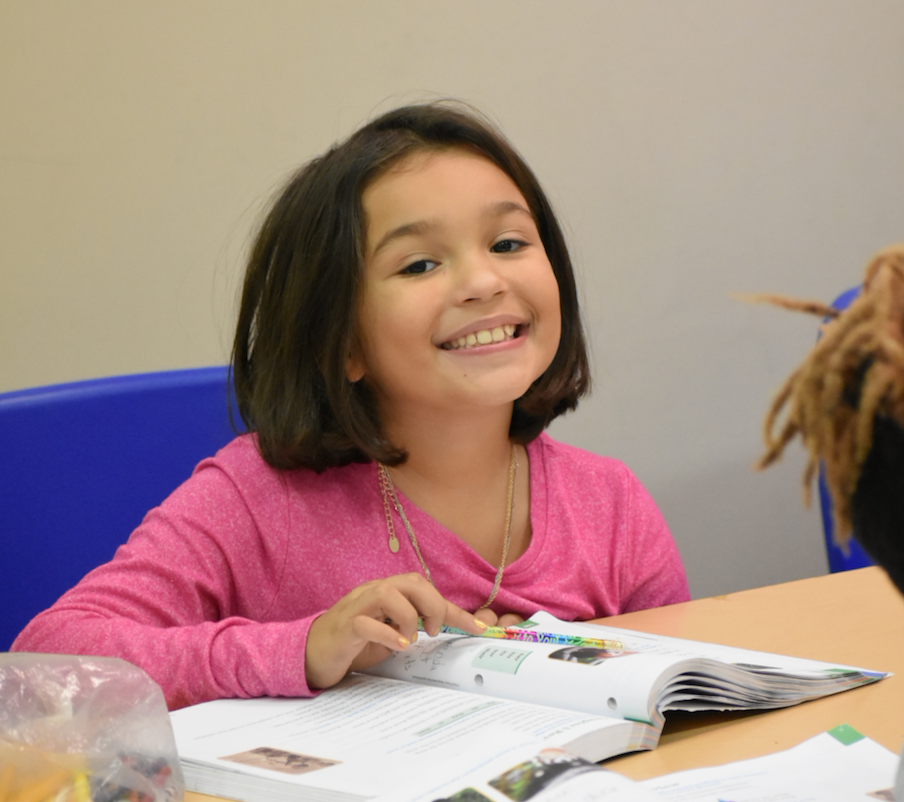 Girl smiles at desk, looking at open book. Pink shirt, short dark hair, blue chair.