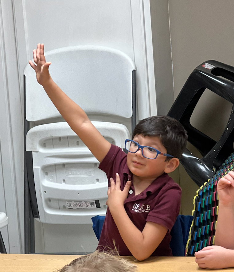 Young boy with glasses, arm raised, hand open, in a classroom setting with a desk.