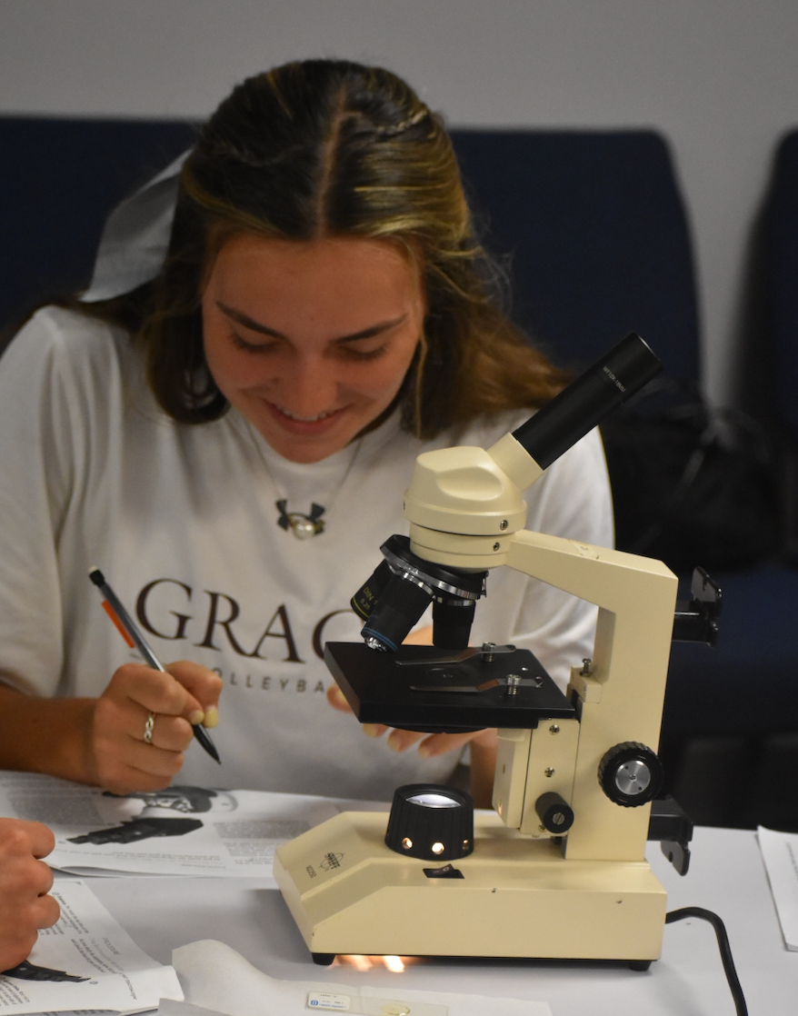 Woman using a microscope, writing notes. She wears a white shirt, is in a lab setting, and appears focused.