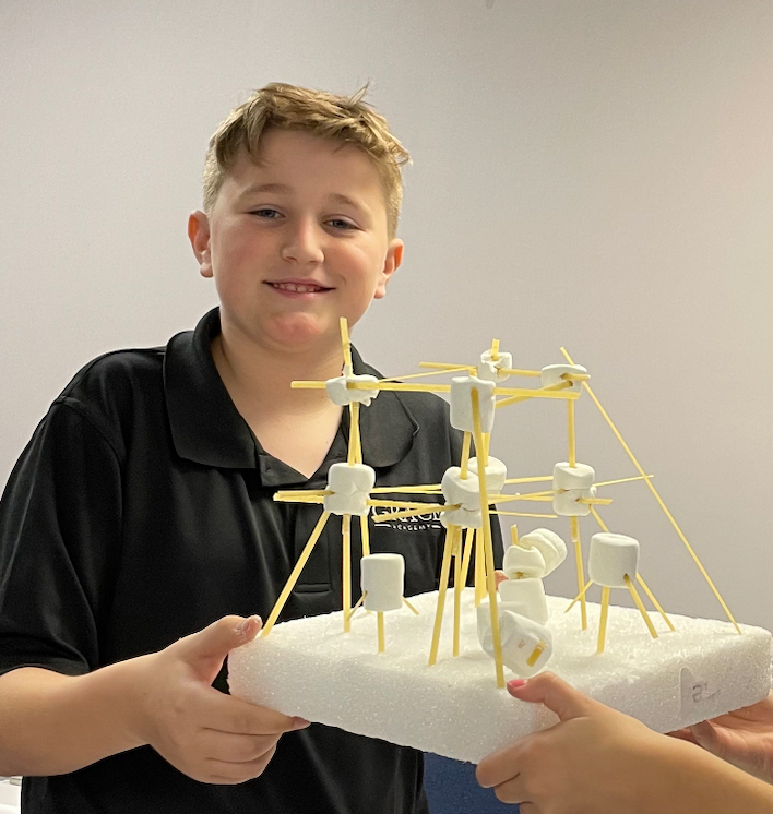 Boy holding a marshmallow and toothpick tower. Smiles. White background.