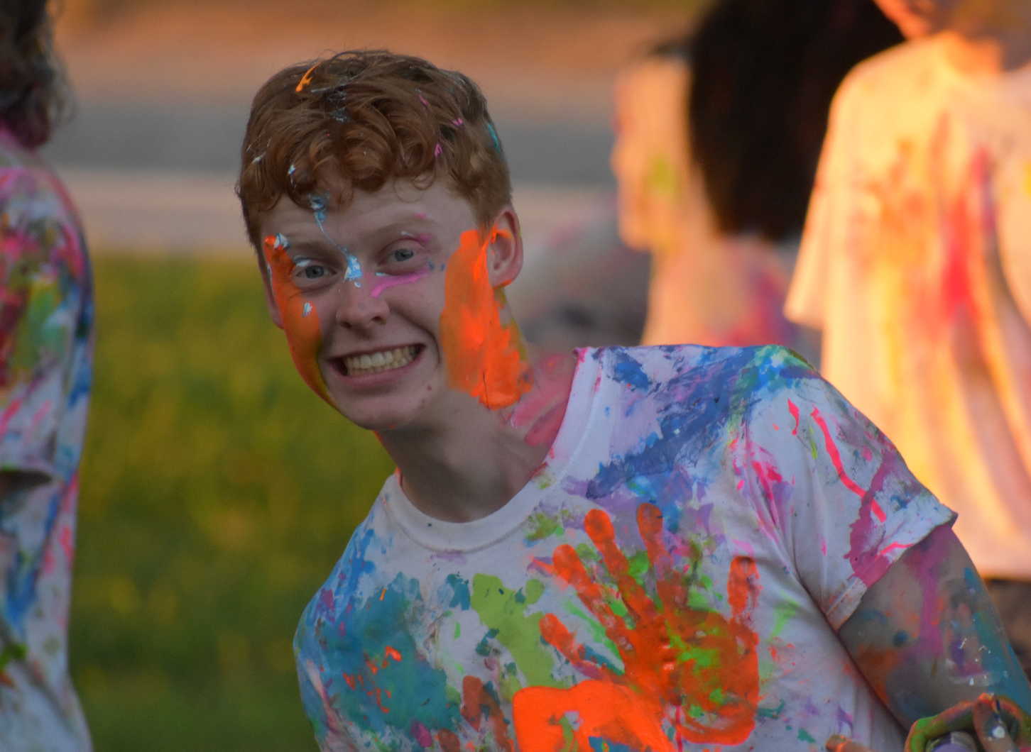 Young person with red hair, covered in colorful paint at a paint party, smiling.