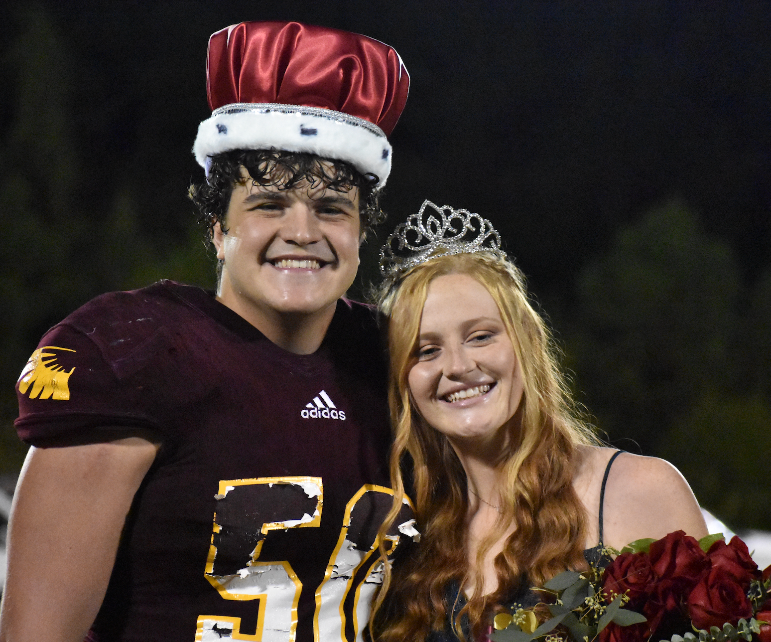 Homecoming king and queen smiling, he in football uniform, she in a gown, both wearing crowns.