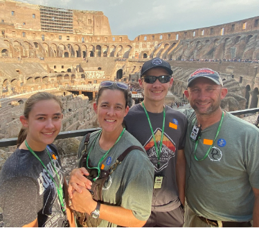 Family posing in front of the Colosseum in Rome. They are smiling, wearing casual clothes and tourist passes.
