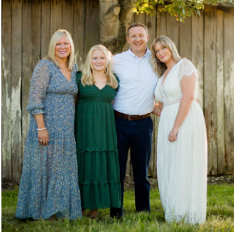 Family of four poses outside. Two women in dresses flank a man in a white shirt. Another woman smiles to the right.