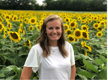 Woman smiling in a field of yellow sunflowers. She wears a white shirt, trees in background.