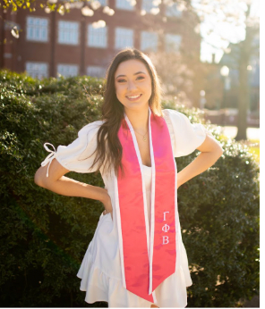 Woman in white dress and pink sash smiles outdoors. Building and bushes in background.