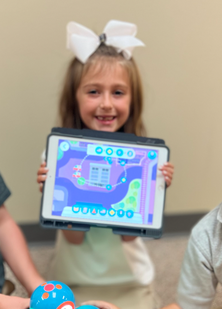 Girl with bow smiling, holding tablet showing a colorful coding game.