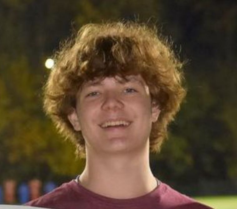 Young man with curly brown hair smiles, wearing a burgundy shirt, outdoors at night.