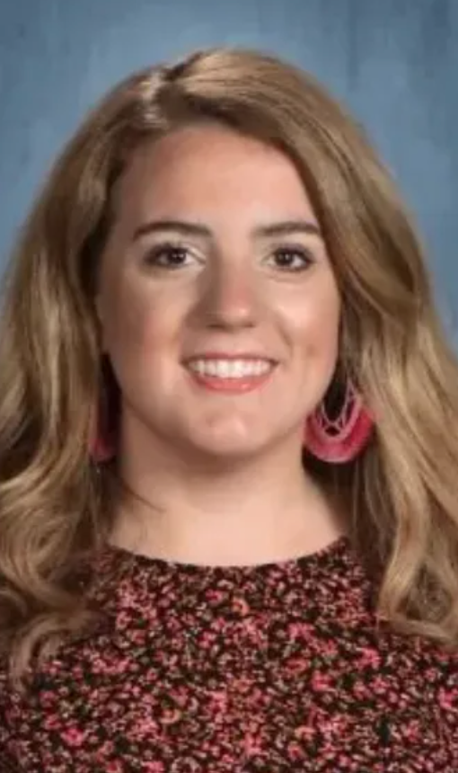 Blonde woman smiles. She wears a floral top and pink earrings against a blue background.