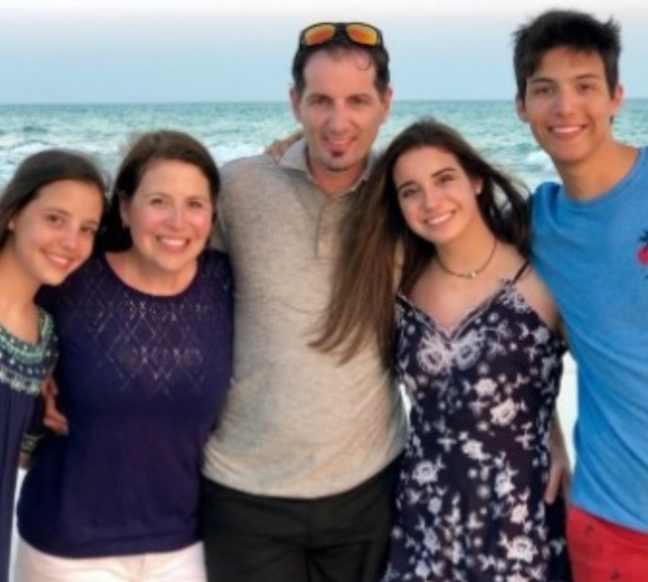 Family of five smiles on a beach. Father with sunglasses, two daughters, and a son embrace at sunset.