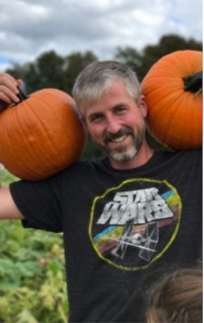 Man with graying hair holding two pumpkins, wearing a Star Wars t-shirt in a pumpkin patch.