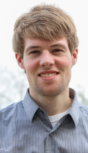 Blond-haired man smiling, wearing a striped button-up shirt, outdoors.