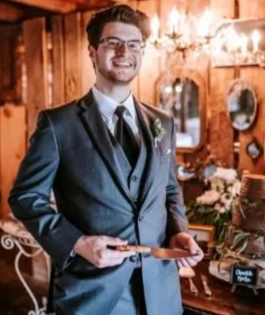 Man in gray suit holding cake server, smiling at wedding.