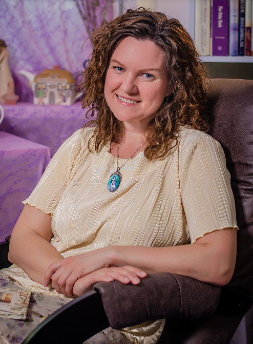 Woman with curly hair smiles, seated in brown chair, wearing cream top, necklace, purple backdrop.