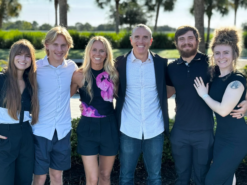 Family of six smiles, arms around each other, outdoors. Sunny day, green grass, palm trees in background.