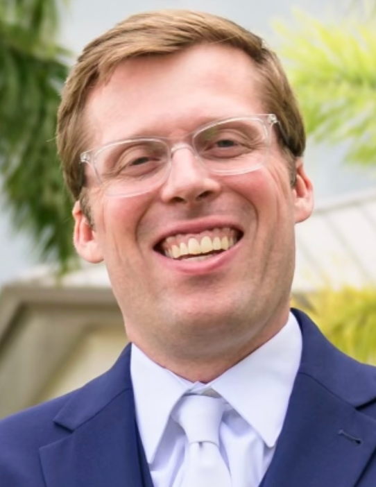 Man in suit and glasses smiles widely outdoors.