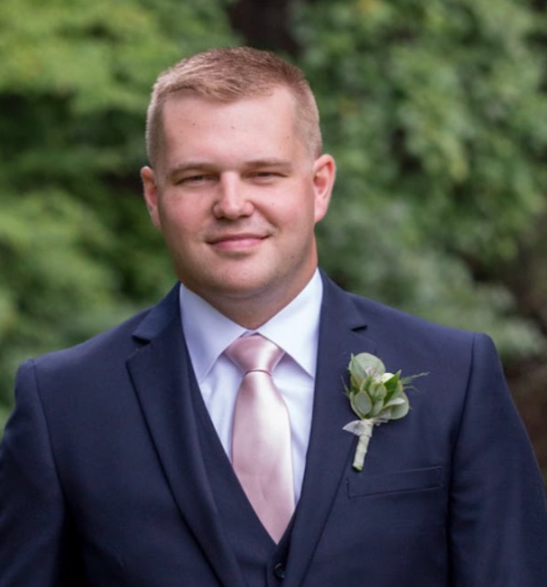 Man in navy suit with pink tie, smiling, against a blurred green background.