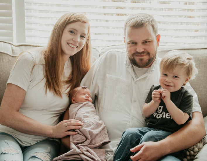 Family of four: parents with newborn and toddler. Woman smiles, holding baby. Man holds toddler.