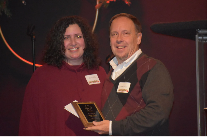 Woman and man at an event, man holding award, both smiling. Maroon outfits, dark background.