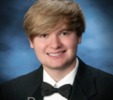 Young man with blonde hair, wearing a black tuxedo and bow tie, smiles at the camera against a blue backdrop.