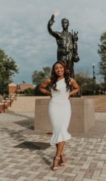 Woman in white dress poses by statue in a brick plaza, smiling.