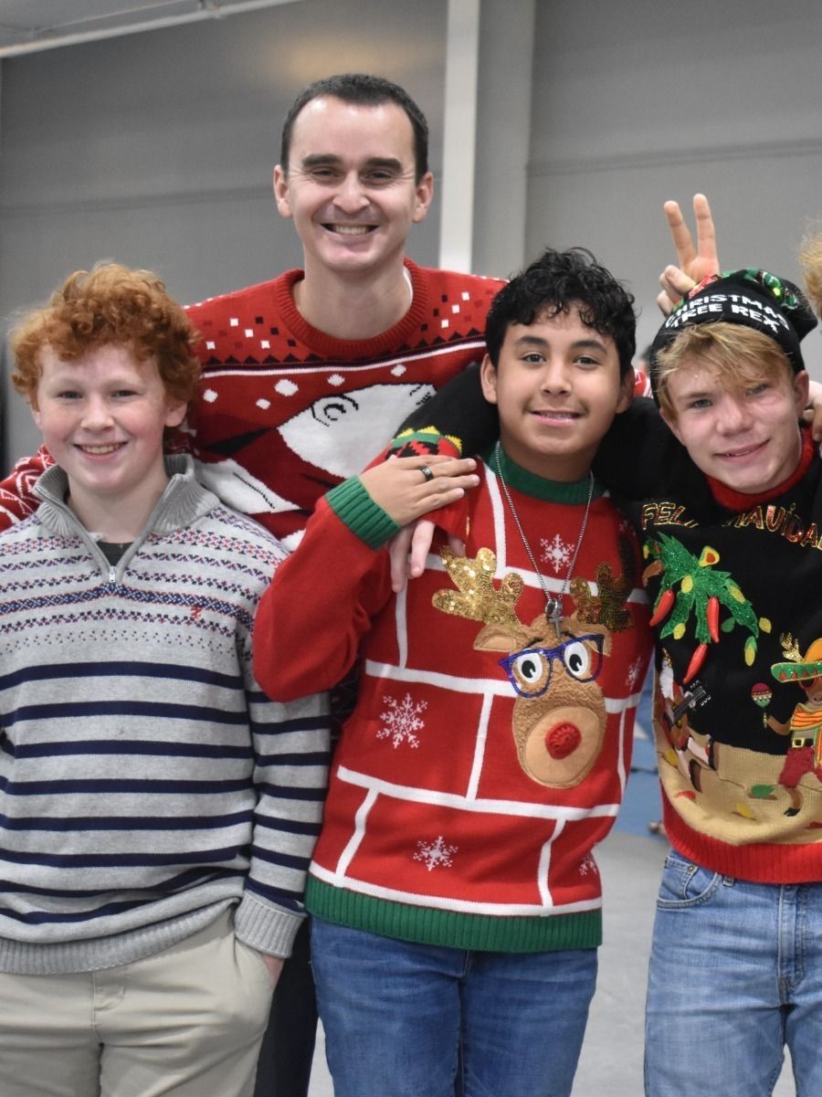 Four people in festive sweaters smile for a photo. The man in back has a holiday sweater.