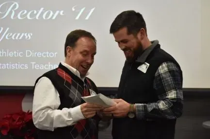 Two men look at a paper, likely a gift. One wears a sweater vest, other a flannel and vest, in a room with a screen.