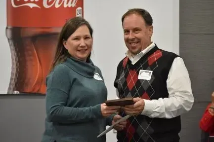 Woman in teal sweater presents a gift to a smiling man wearing a sweater vest in front of a Coca-Cola backdrop.