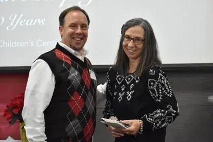Man and woman standing side-by-side; man in argyle vest, woman holding a gift.