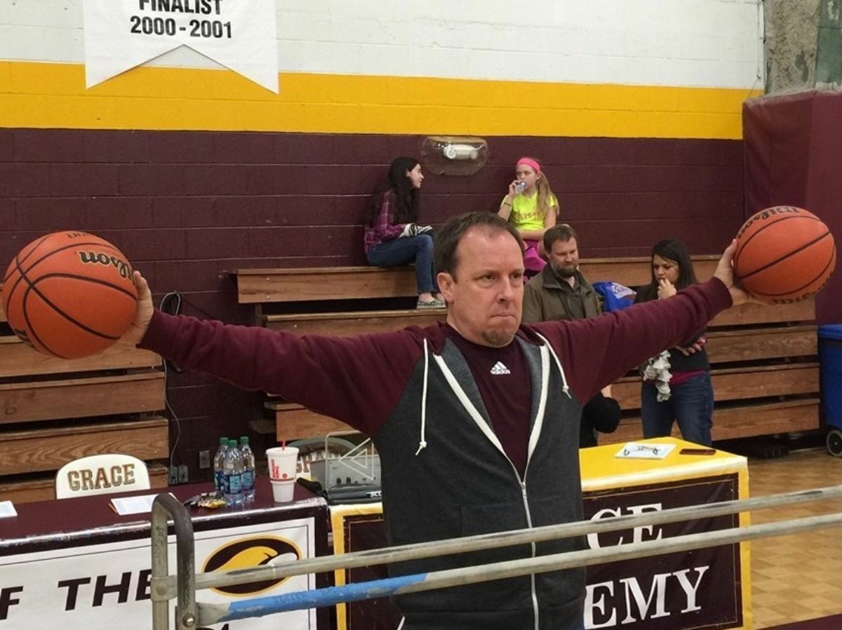 Man holding basketballs, arms outstretched in a gymnasium; other people and a banner in the background.