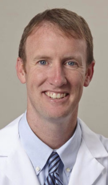 Man in white lab coat smiling, blue eyes, fair skin, wearing a striped tie.