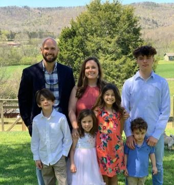 Family posing outdoors with a backdrop of mountains and trees. Smiling, includes two parents and four children.