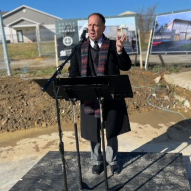 Man in suit speaking at a podium outdoors, gesturing with a hand; construction site in background.