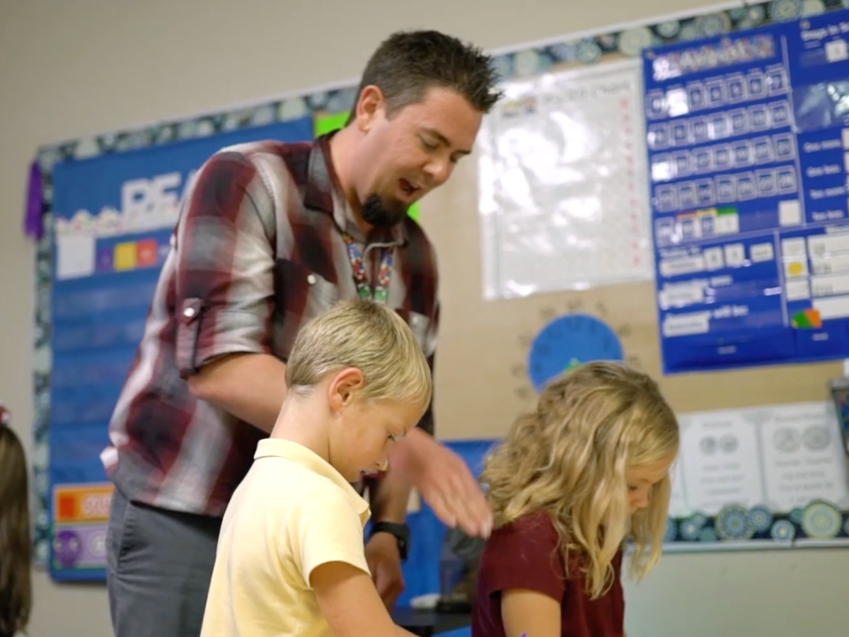 Teacher in plaid shirt assisting two young students at their desks. Classroom setting.