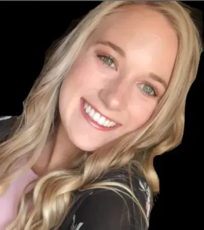 Blonde woman with a bright smile, wearing a pink shirt and black jacket, posing for a photo.