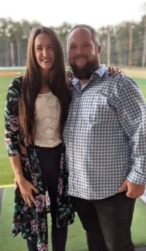Woman with long brown hair and floral cover-up poses with a man with a beard, outdoors.