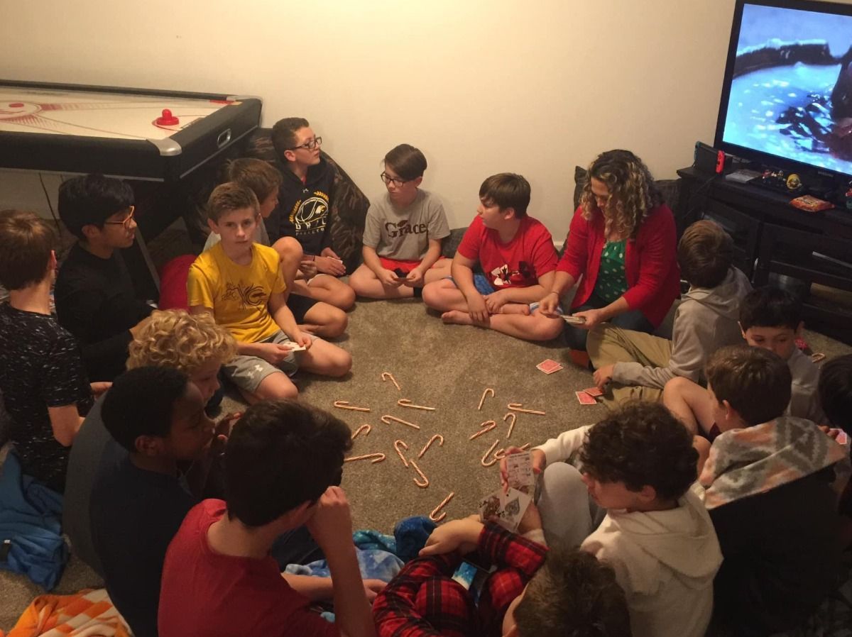 A group of kids and adults sit in a circle, playing a game with sticks indoors.