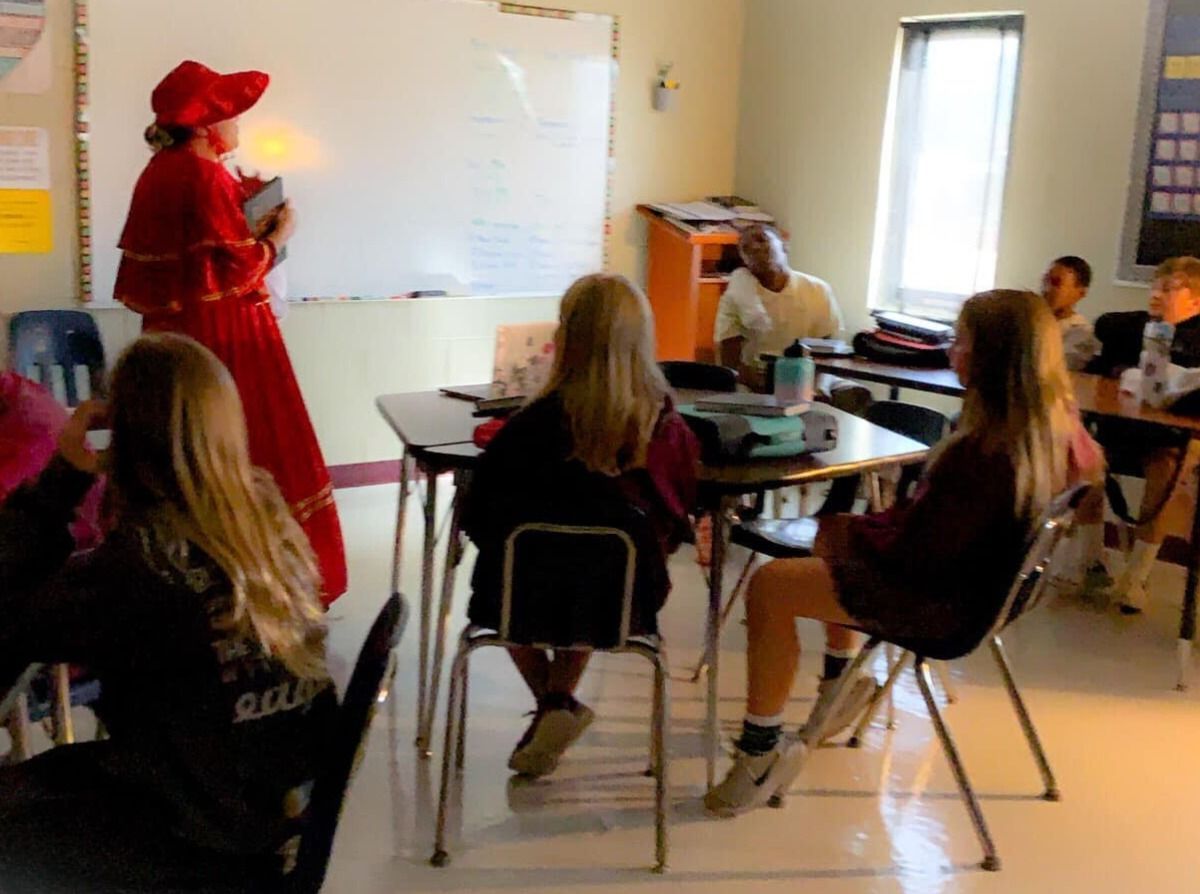 A costumed person lectures students in a classroom. The teacher wears red and holds a book near a whiteboard.