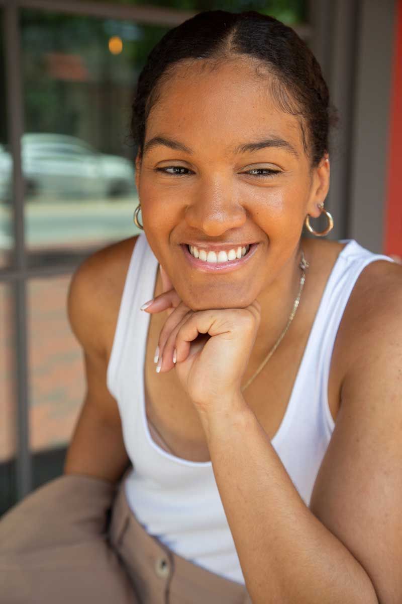 A woman in a white tank top is smiling with her hand on her chin.