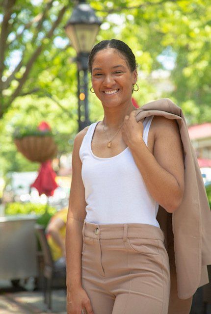 A woman in a white tank top and tan pants is holding a jacket over her shoulder.