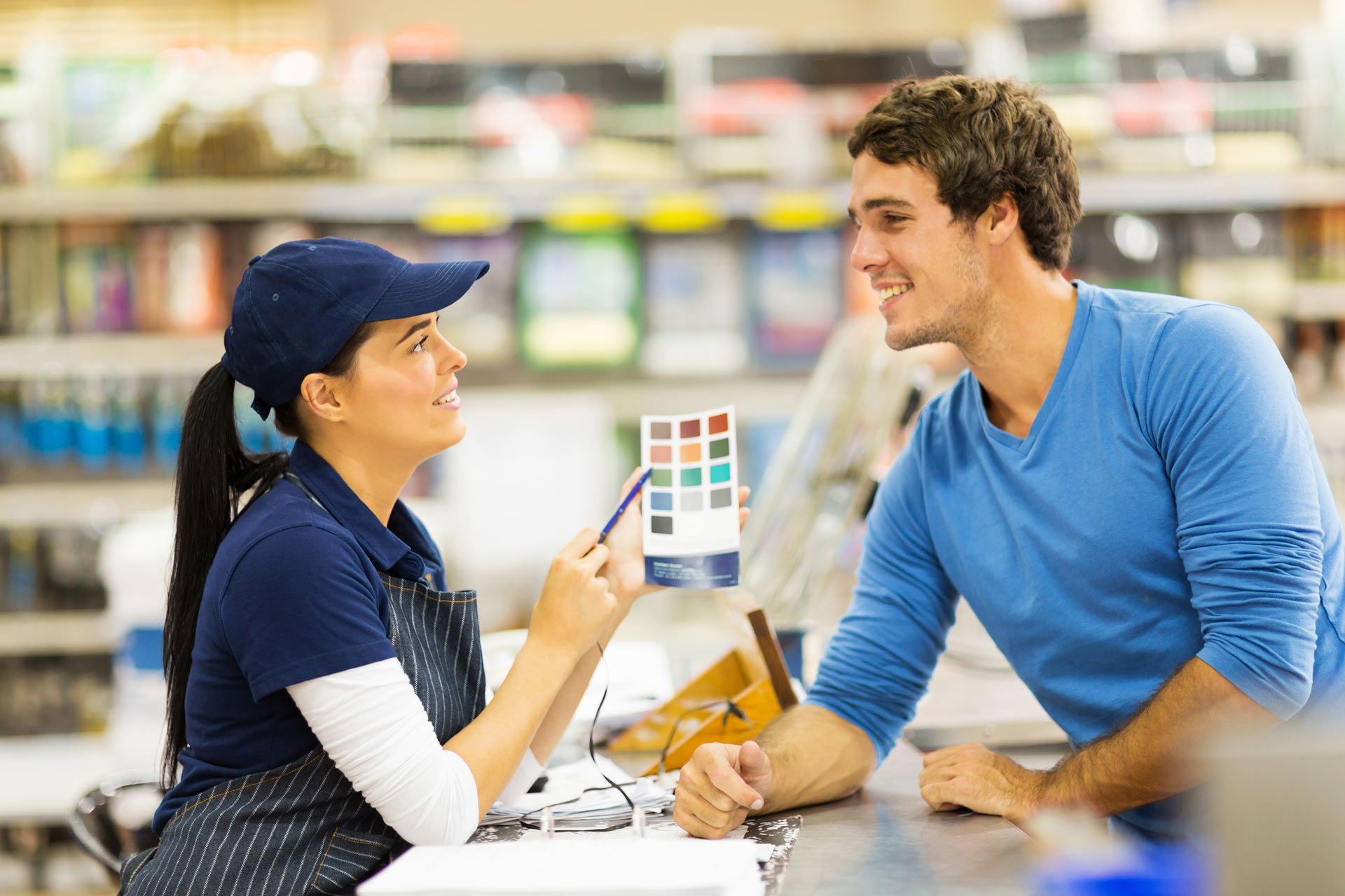 Man & woman discuss art supplies in a painting store, with various paintings displayed around them.
