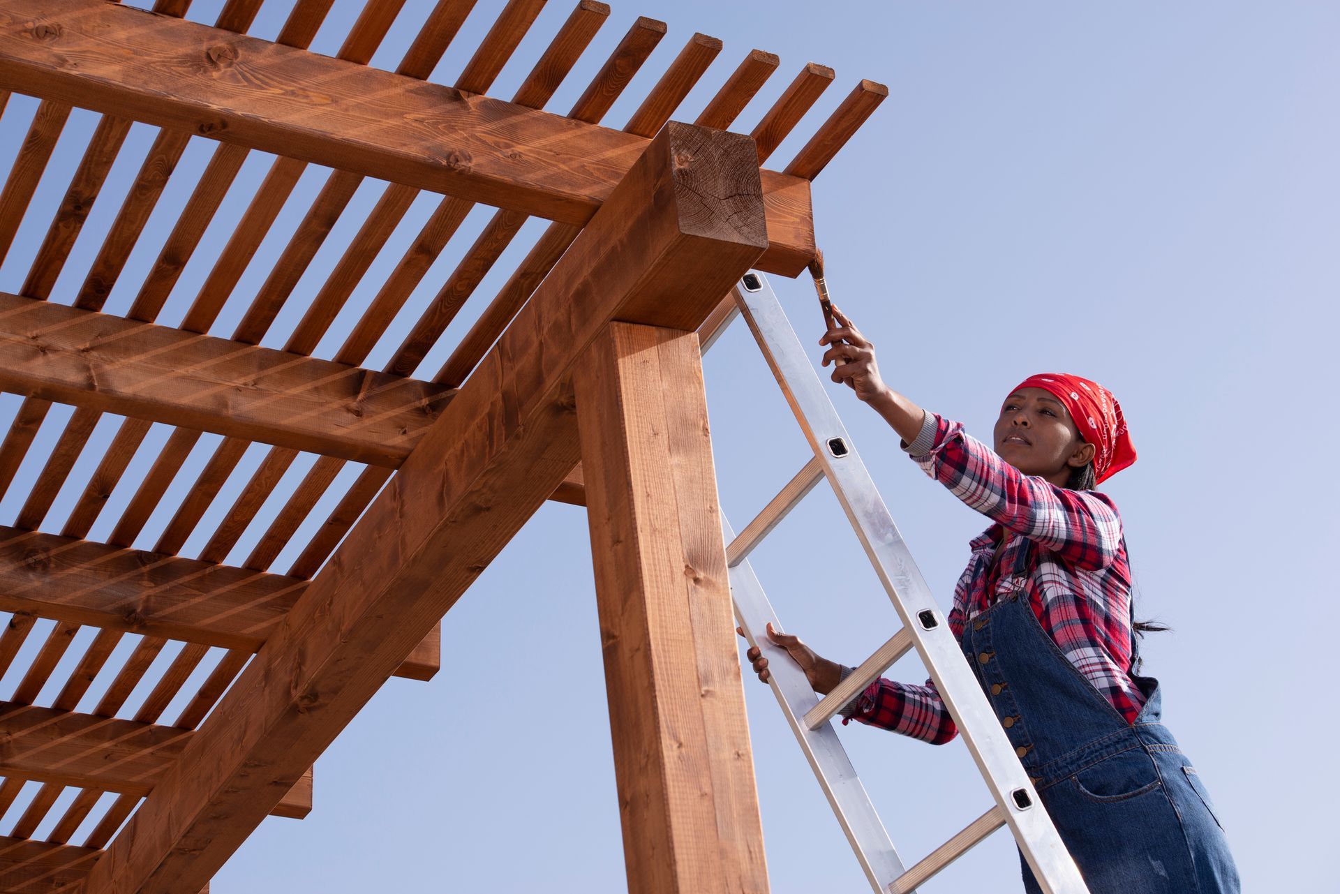 A woman is standing on a ladder painting a wooden structure.