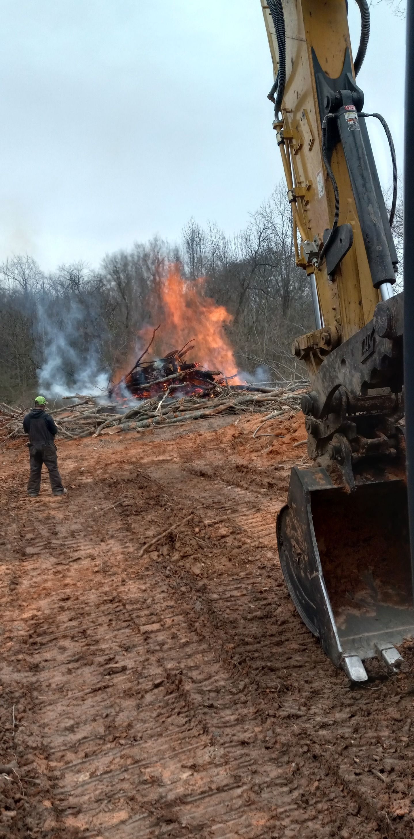 A man is standing in the dirt next to a bulldozer with a fire in the background.