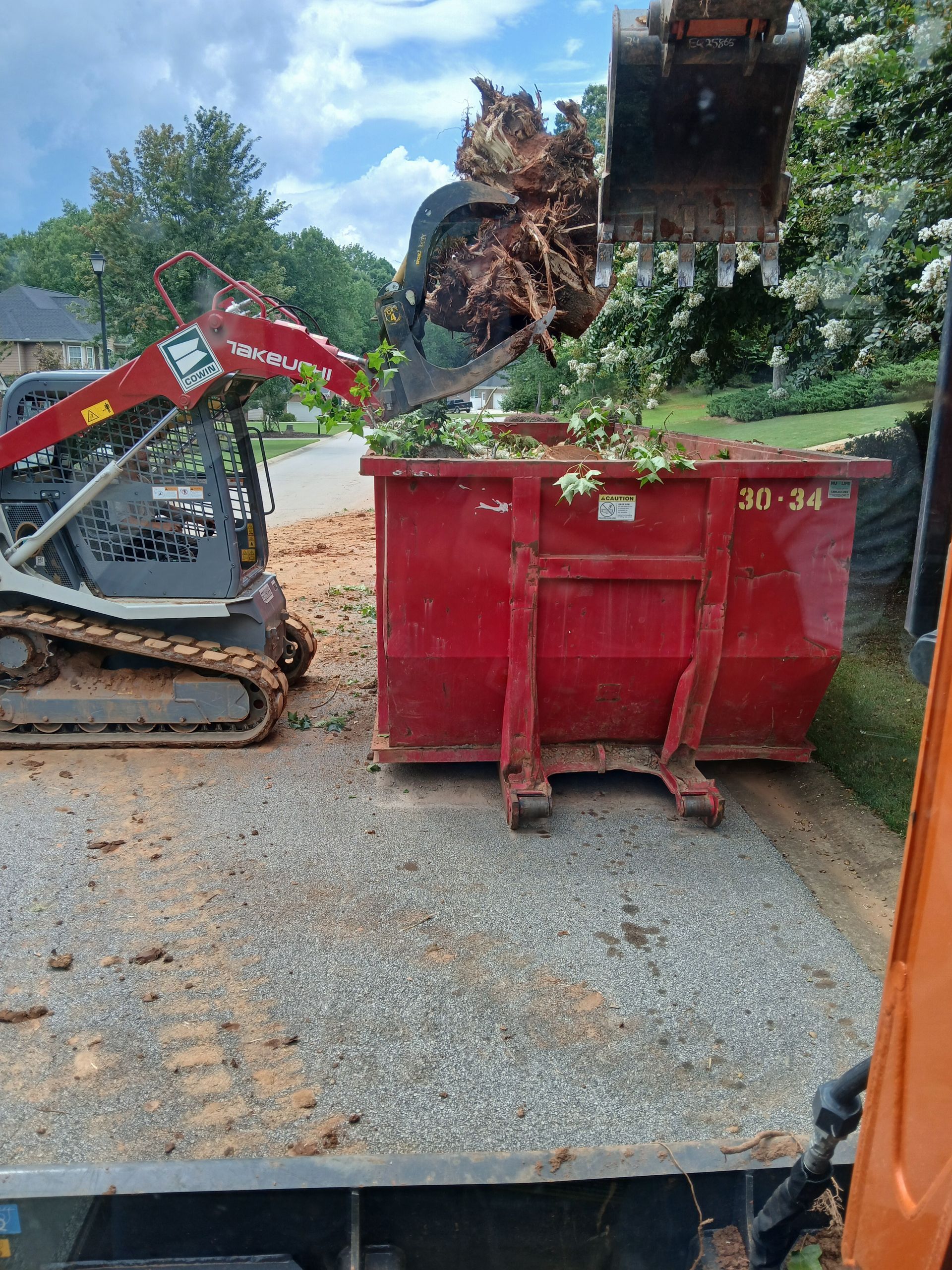 A bulldozer is loading a red dumpster with a tree stump in it.