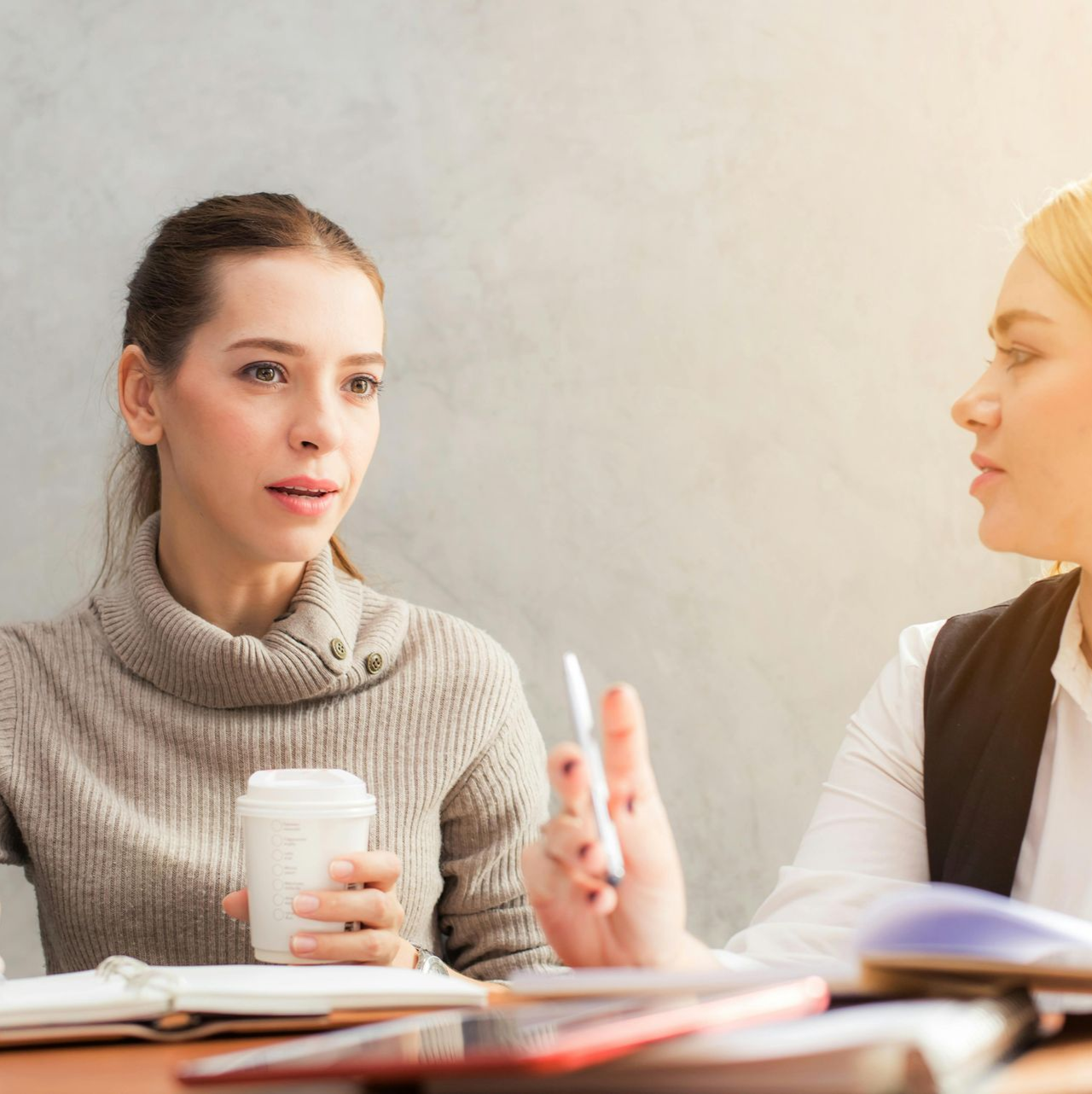 שתי נשים ליד שולחן דנות במסמכים, אחת מחזיקה כוס קפה, השנייה עט.