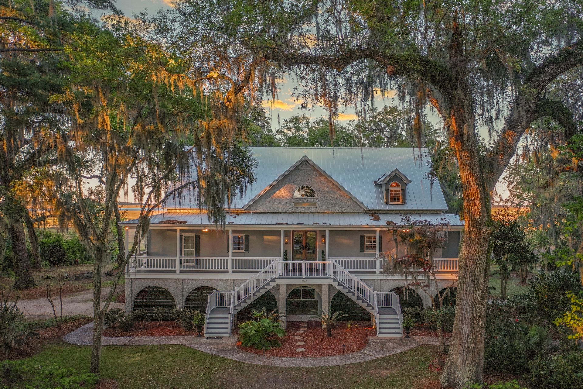Two-story house with wrap-around porch, metal roof, and large trees with Spanish moss; sunset in background.