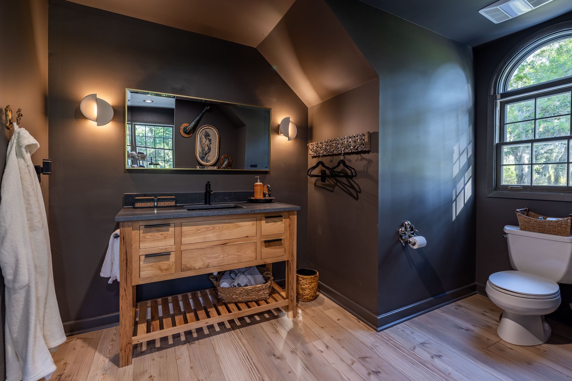 Bathroom with dark gray walls, light wood vanity, arched window, and wooden floor.