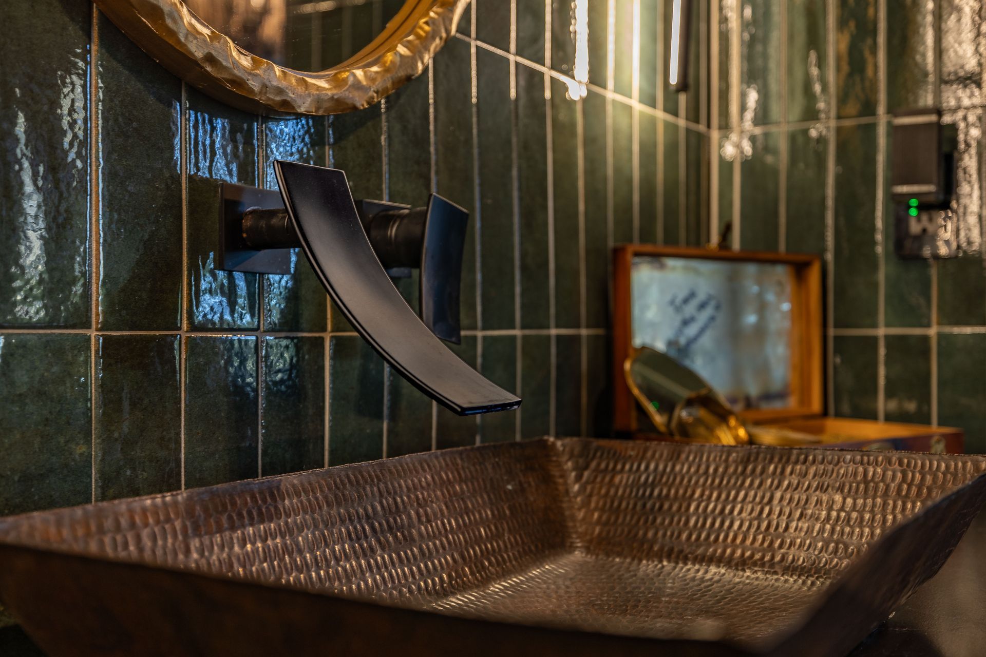 Copper sink with waterfall faucet, green tile backsplash, and decorative mirror in a bathroom setting.