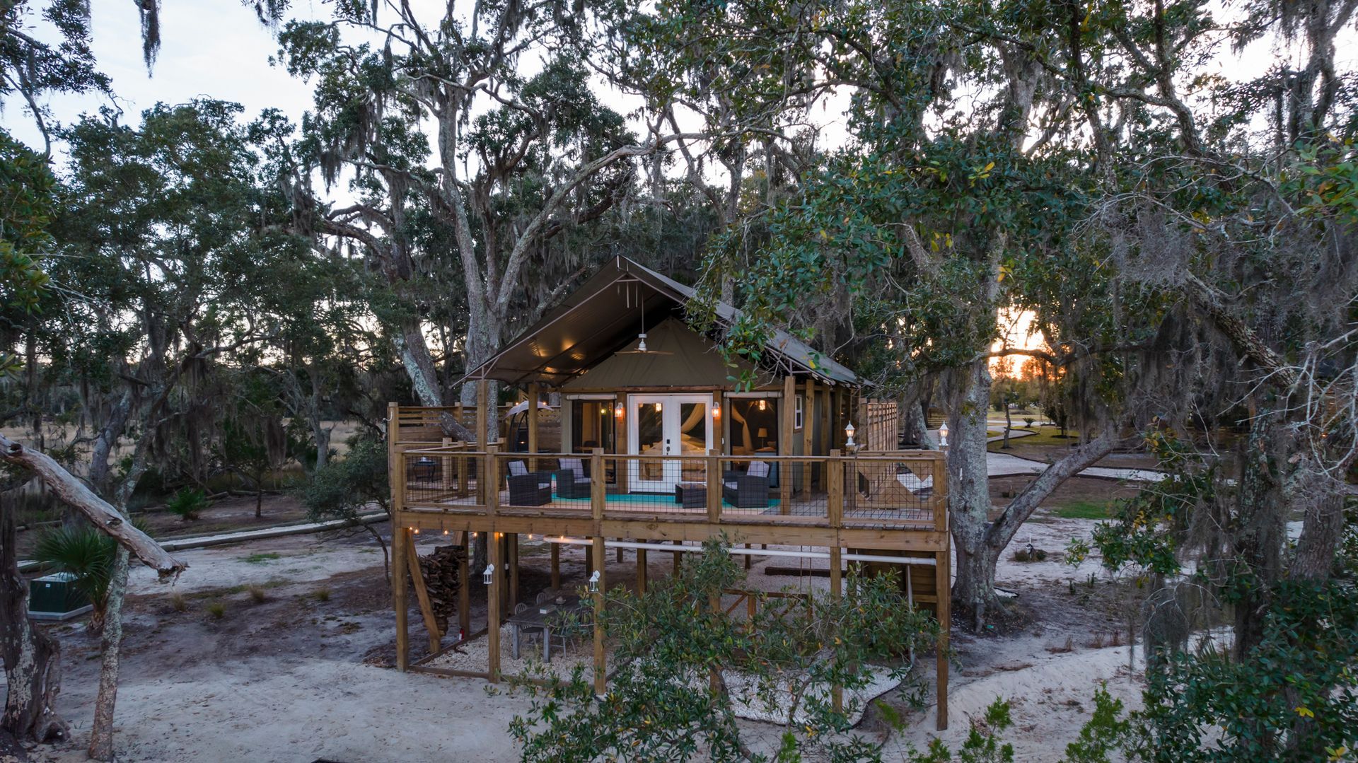 A treehouse with a deck surrounded by trees at sunset; lights twinkle from the deck.